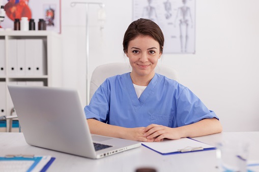 Altura Learning Portrait of medical practitioner nurse looking into camera smiling sitting at desk in conference meeting room. Confident physician asisstant analyzing pills treatment expertise on laptop computer